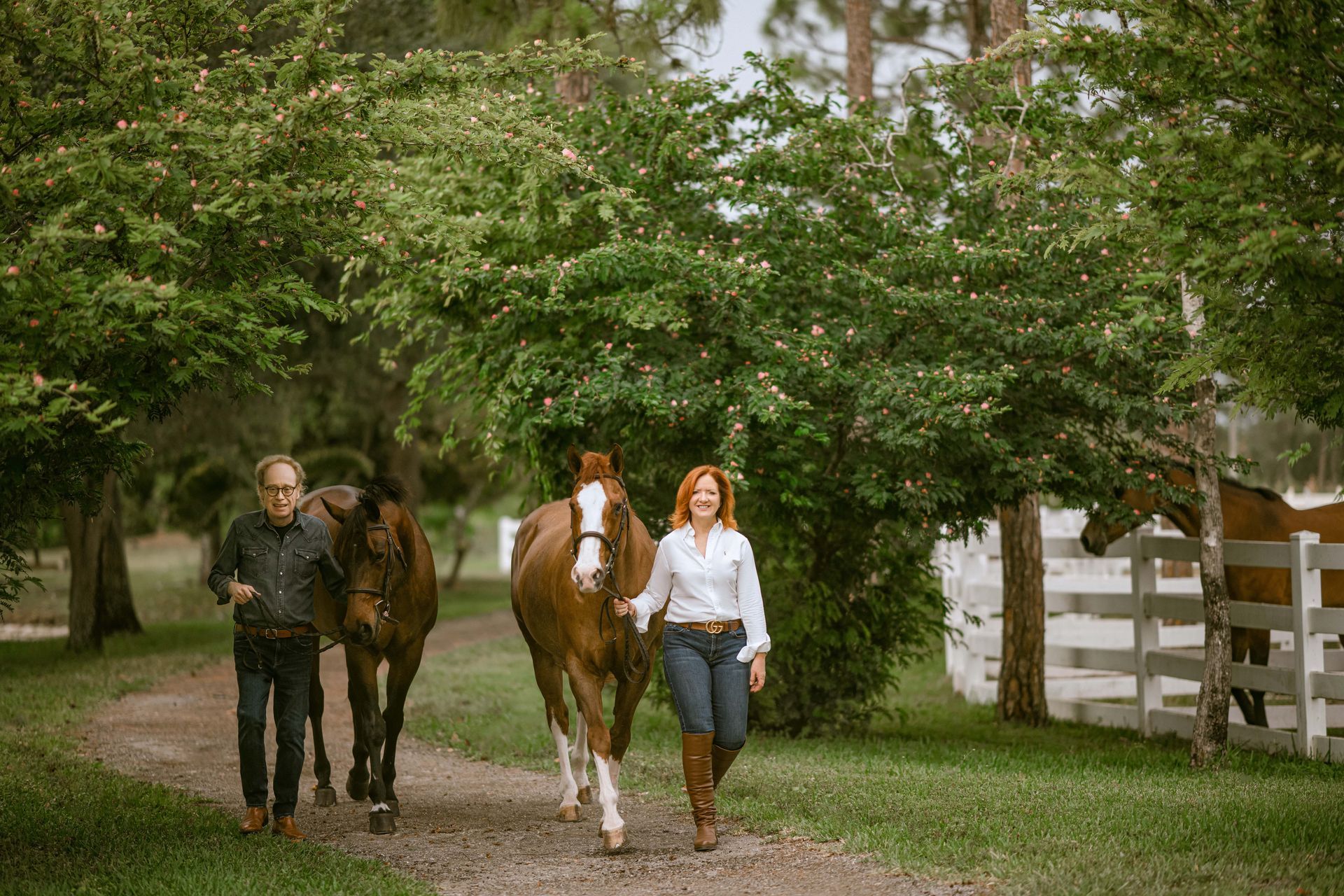 A man and woman walk with two horses on a dirt path. Woman smiles, holding horse. Green trees in background.