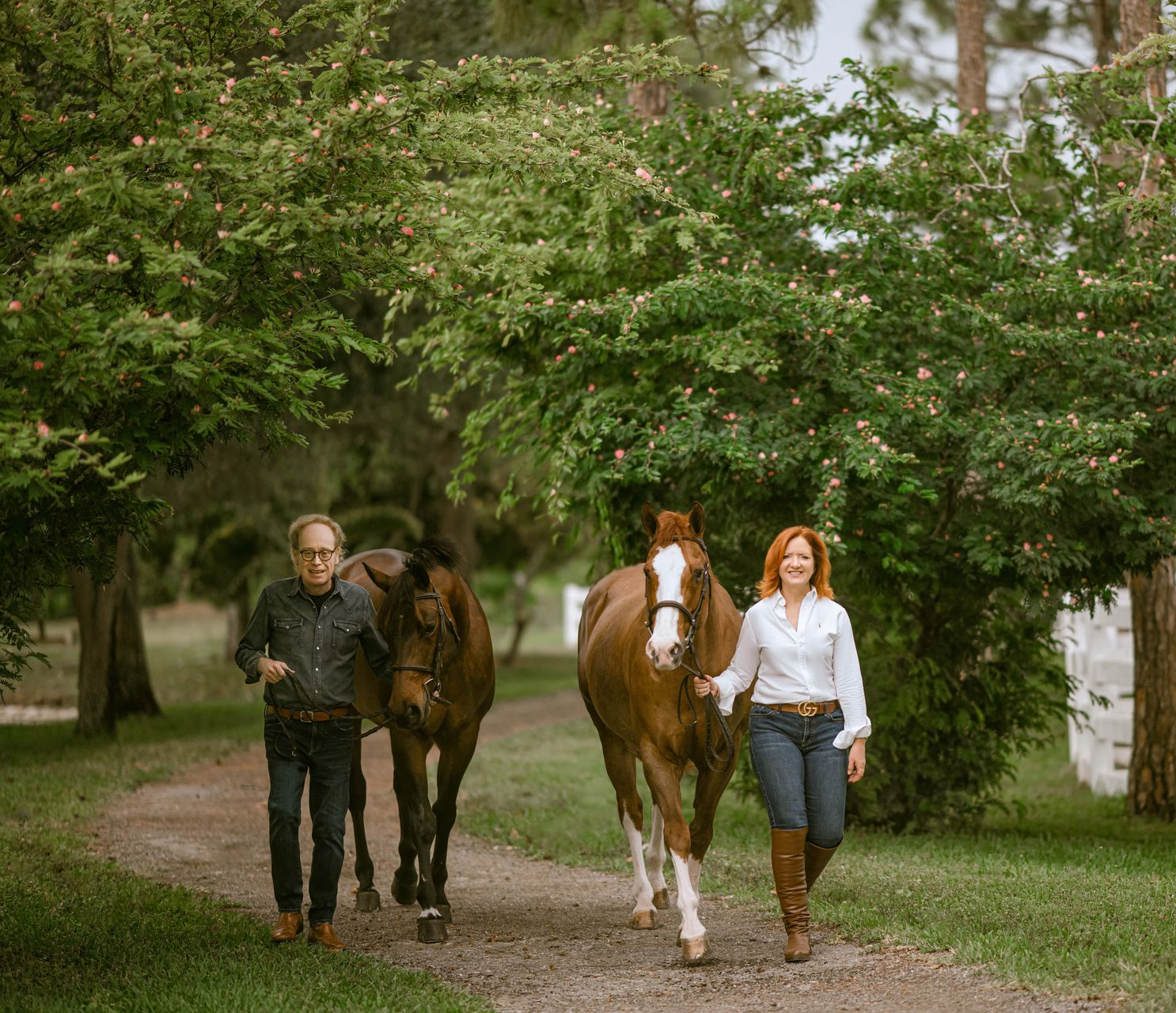 Two people lead horses on a path lined with green trees.