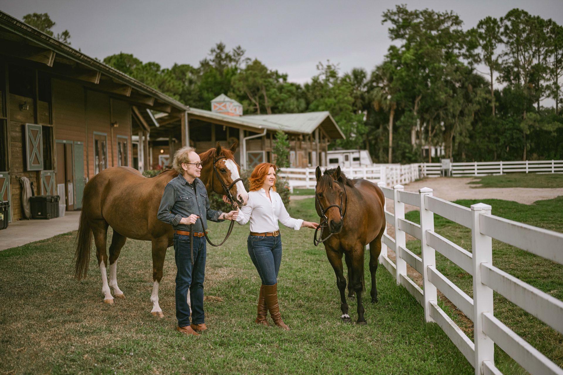 Two women with horses near a barn. One woman touches a horse's head. They stand by a white fence on the grass.