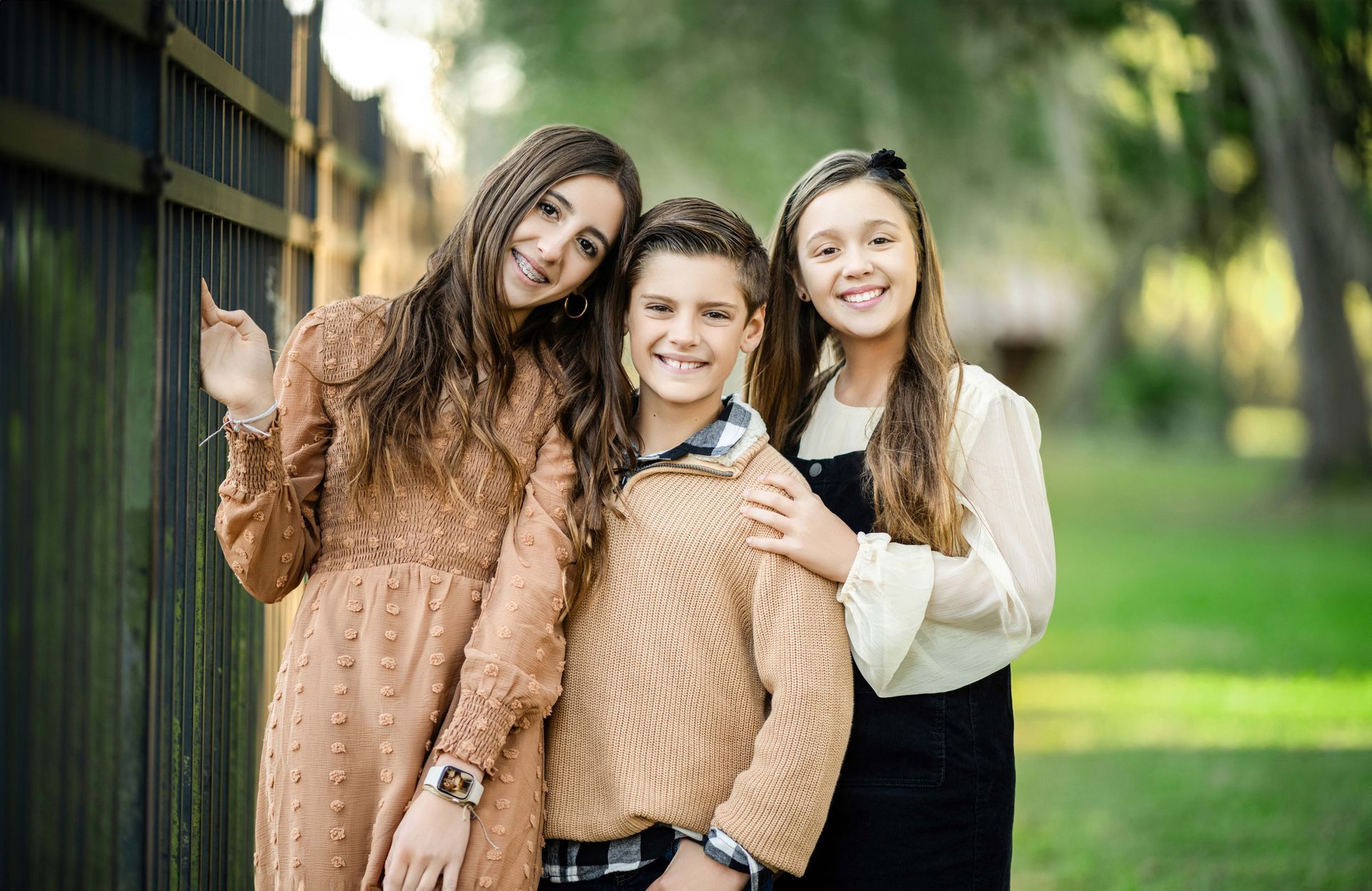 Three smiling siblings pose outdoors. The boy is in the middle, flanked by two girls. Brown and beige clothing, green background.