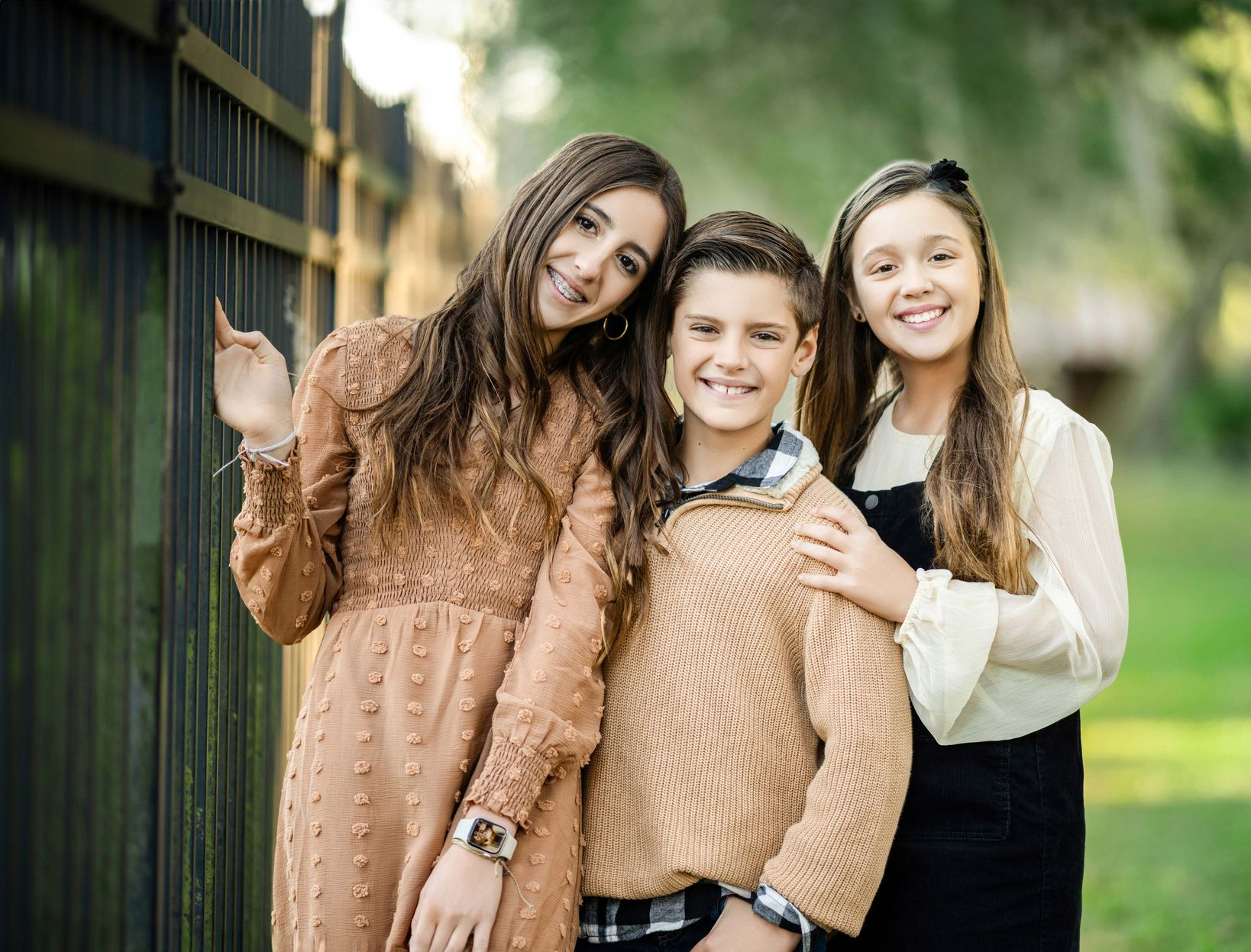 Three smiling children pose near a fence in a park: a girl with long brown hair, a boy in a sweater, and a girl in overalls.