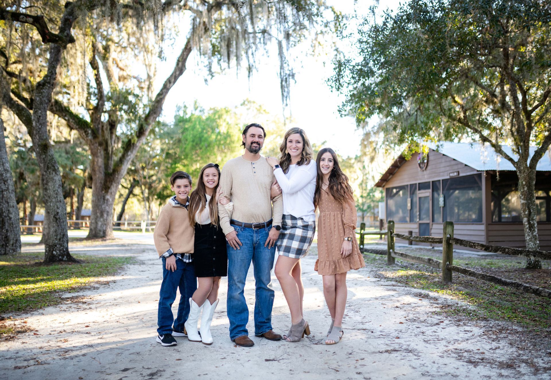 Family of five poses on a path in a park, with a cabin in the background.