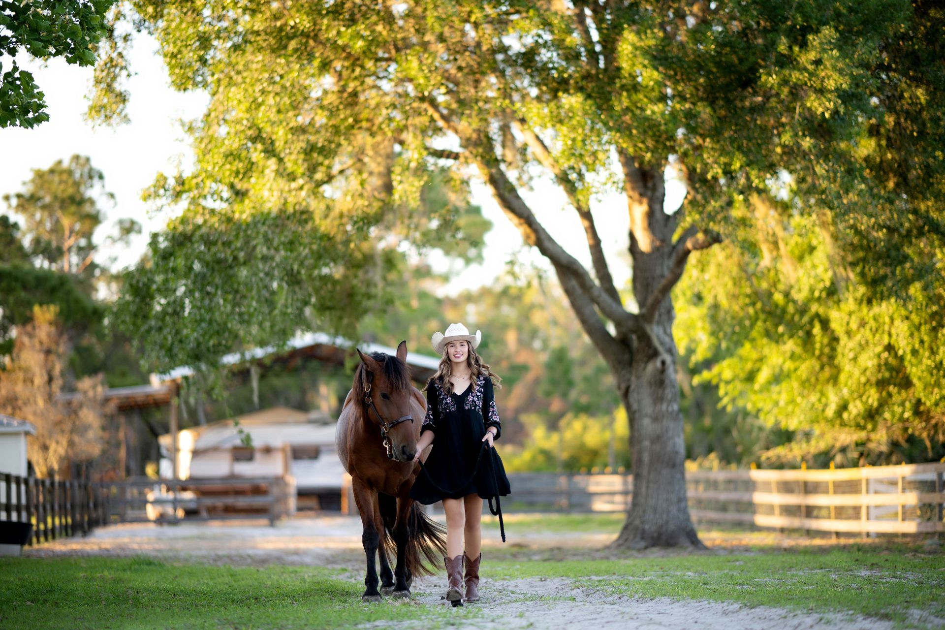 Cowgirl in a black dress, brown boots, and hat, walking a brown horse down a path on a sunny farm.
