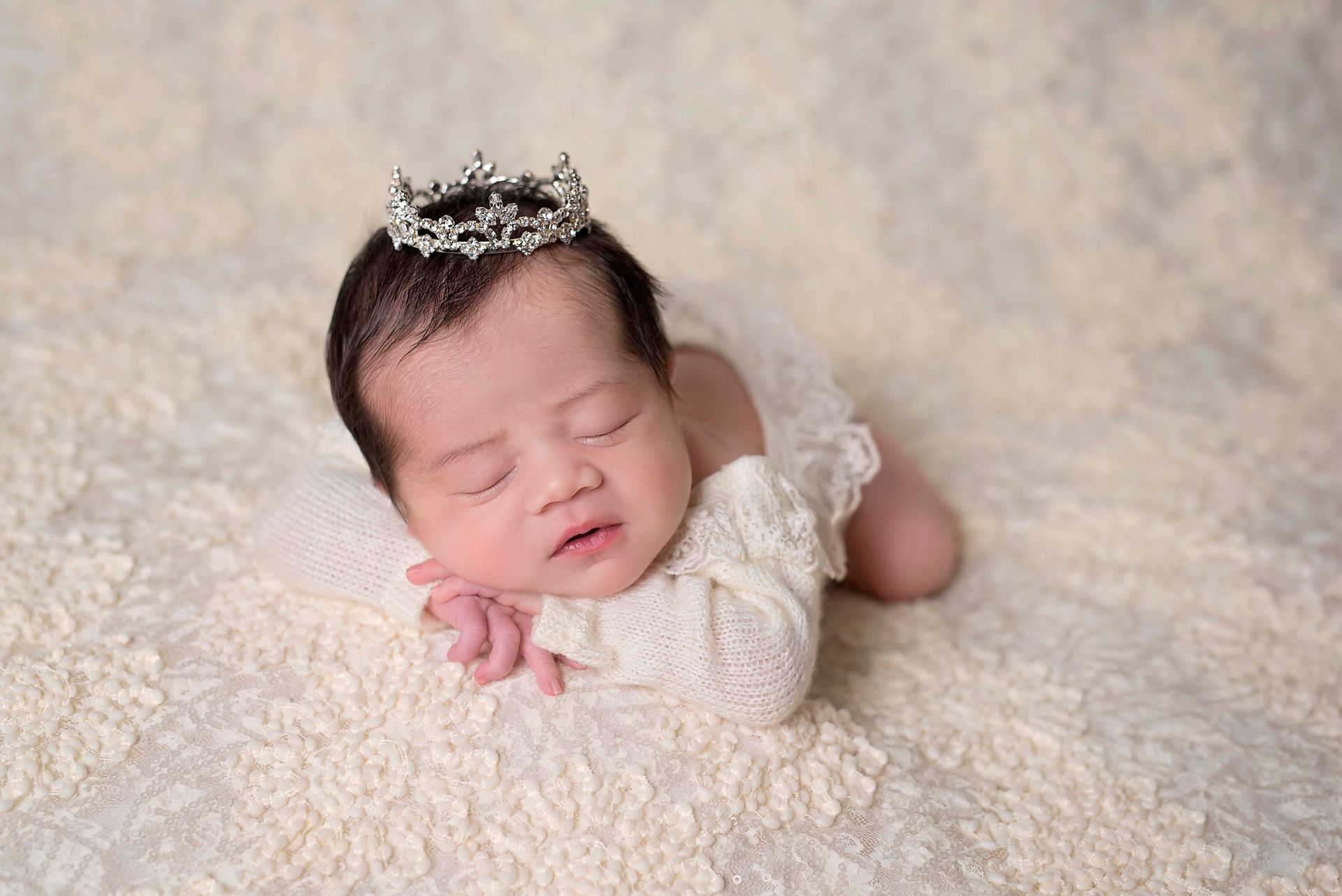 Newborn baby wearing a crown, lying on a cream-colored textured surface, eyes closed.