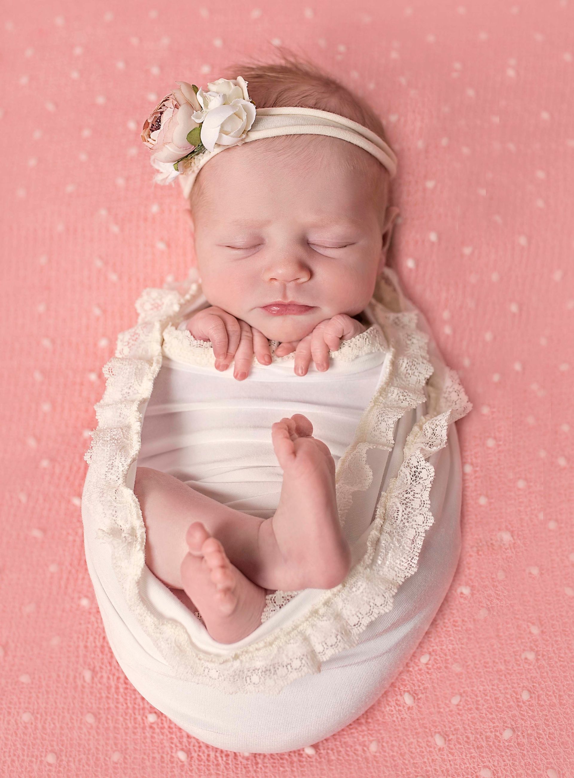 Newborn baby swaddled in white with lace trim, wearing a floral headband, sleeping on pink fabric.