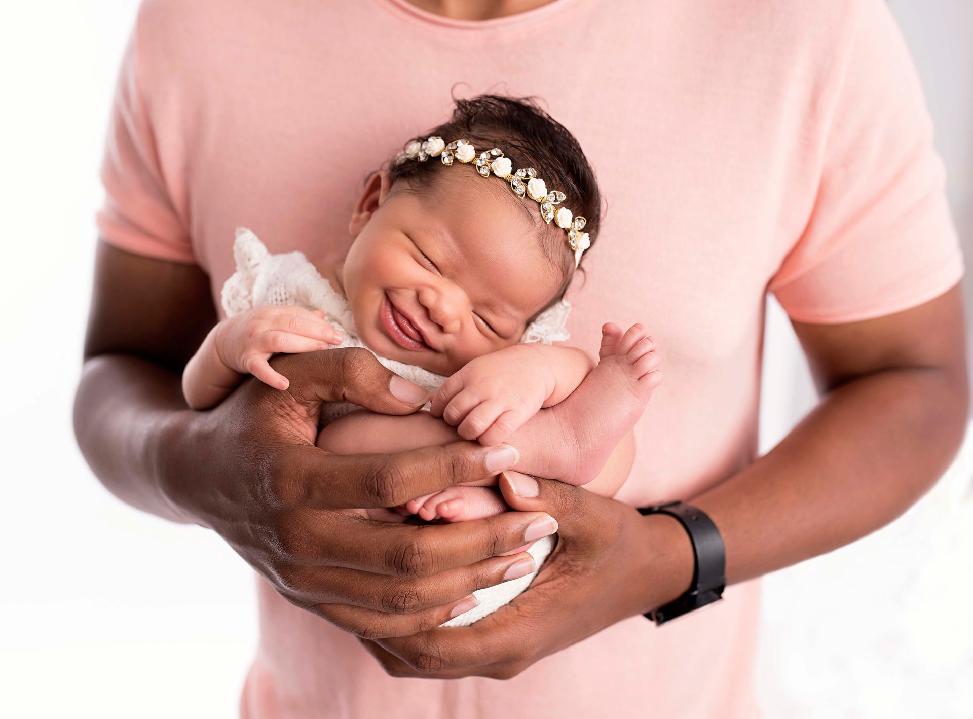 Newborn baby smiling, held by a man in a pink shirt. Baby wears a floral headband.