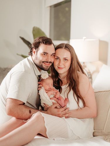 Parents smiling, holding newborn baby. Light-filled room. All wear white, family portrait.