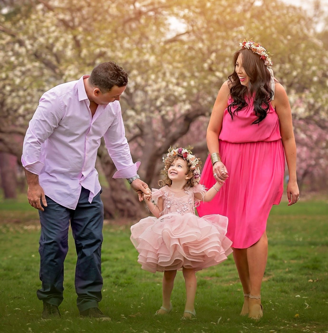 Bride and groom walk hand-in-hand, smiling, under purple wisteria blooms, in a garden setting.