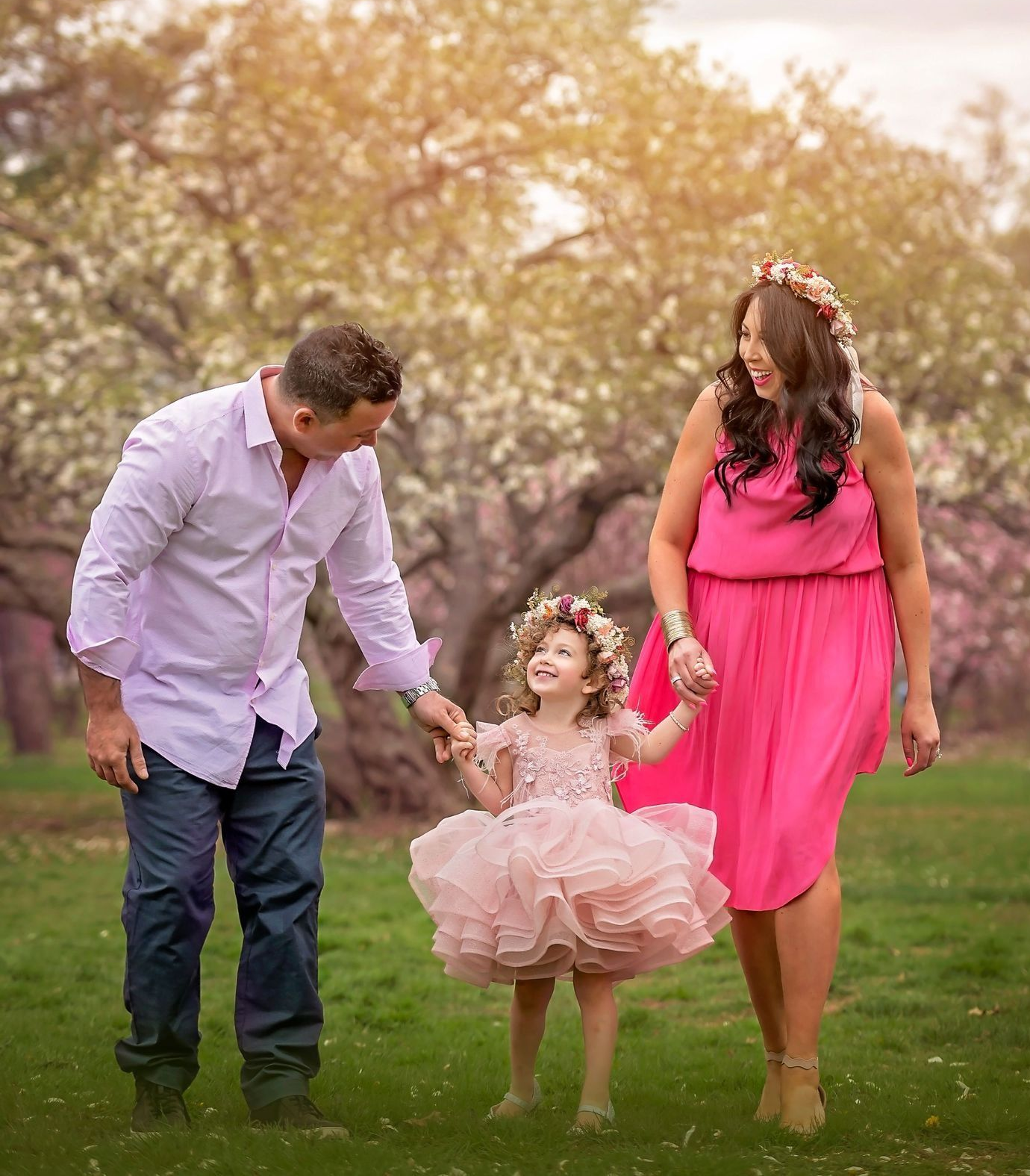Family of three in pink clothing holding hands, walking in a park with blossoming trees.