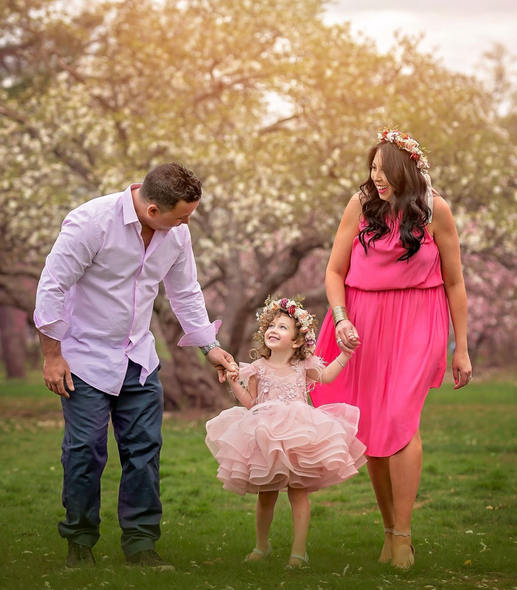 Family of three in pink clothing holding hands, walking in a park with blossoming trees.