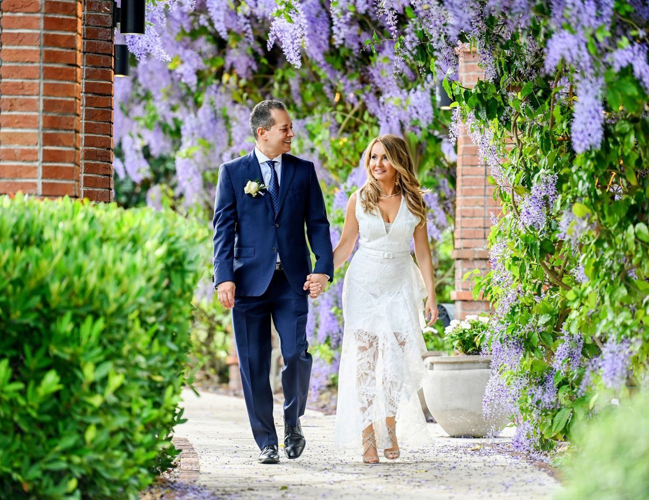 Bride and groom walk hand-in-hand, smiling, under purple wisteria blooms, in a garden setting.