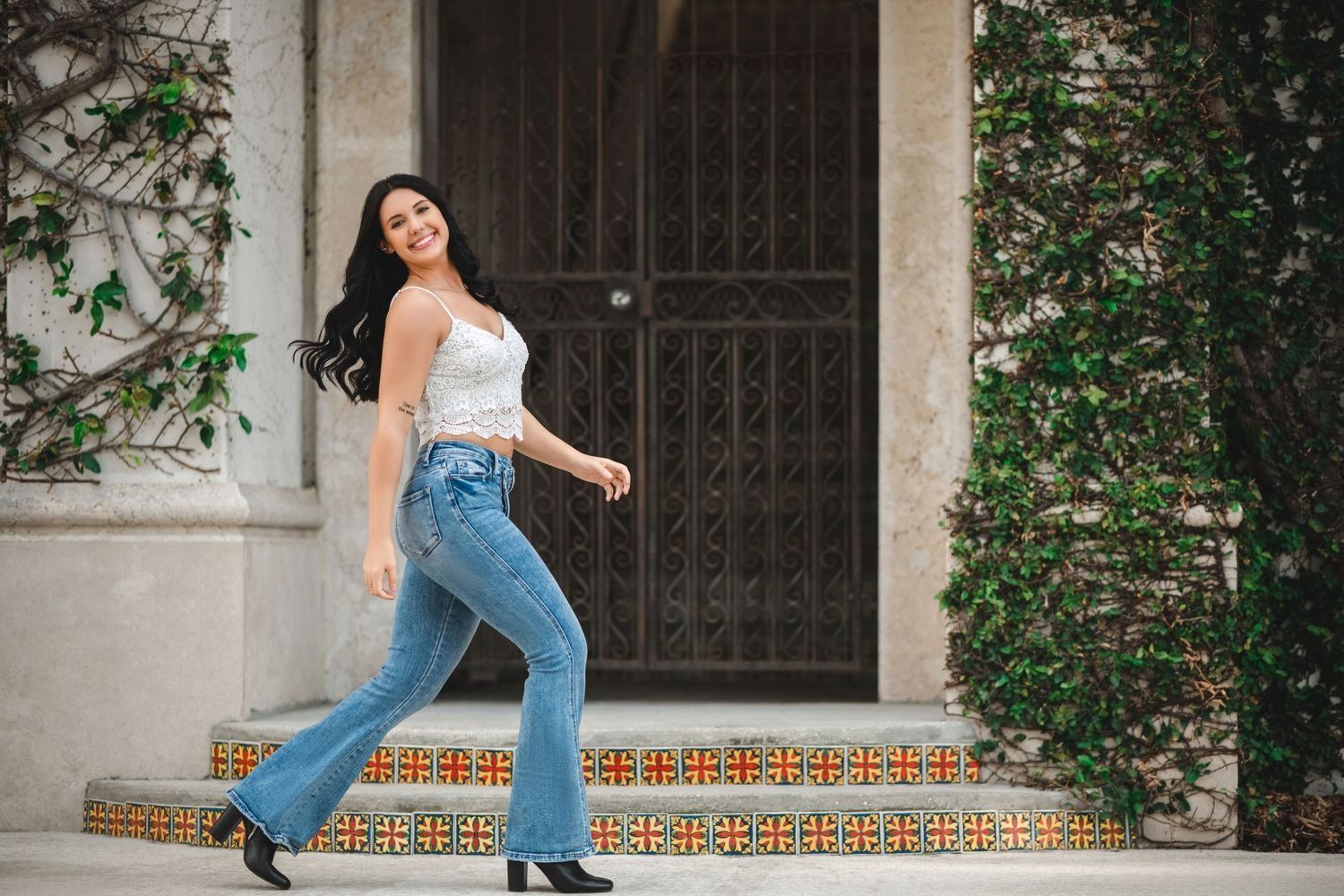 Woman in white top and flared jeans walks up steps; smiling, against a building with a wrought iron door.
