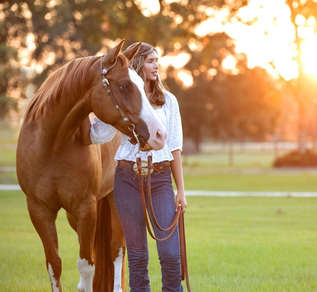 Woman with a chestnut horse in a sunny field, wearing jeans and a white shirt.