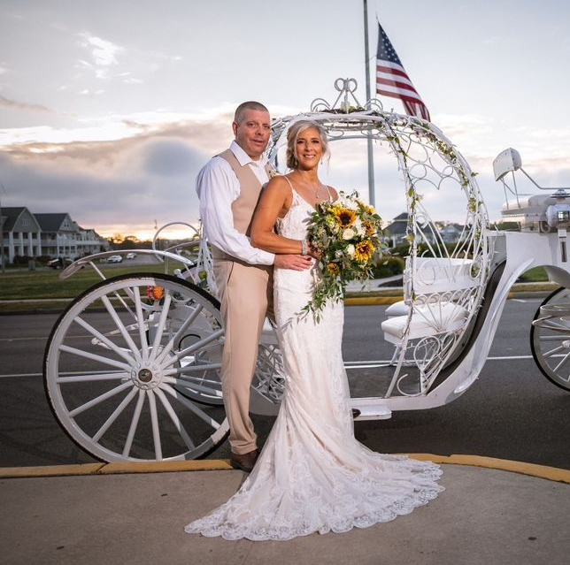 Newlyweds pose beside a white carriage with sunflowers. The man wears tan, the woman, a white gown. American flag flies.