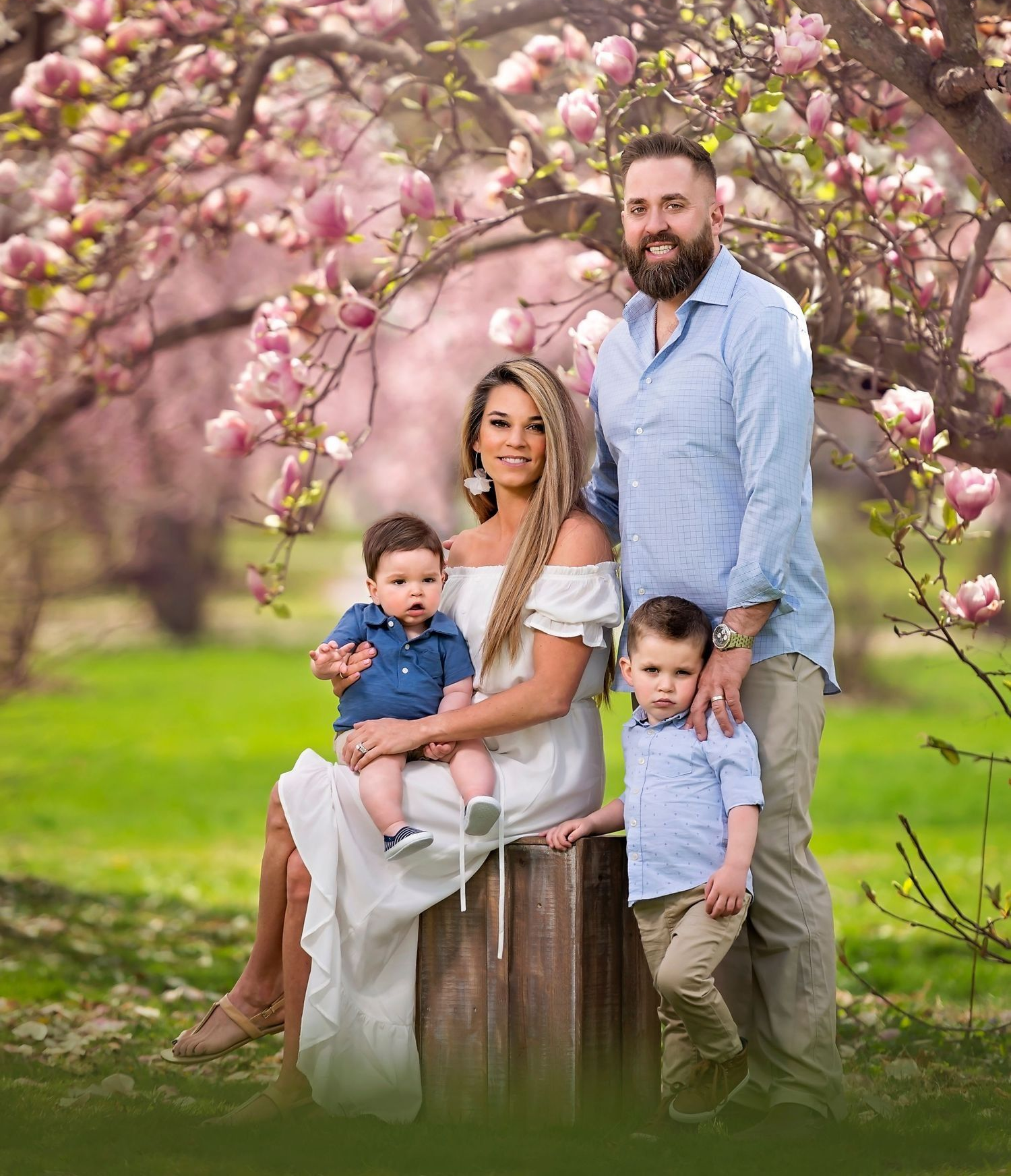 Family of four poses under a flowering tree. Parents and two young boys smile in a park.