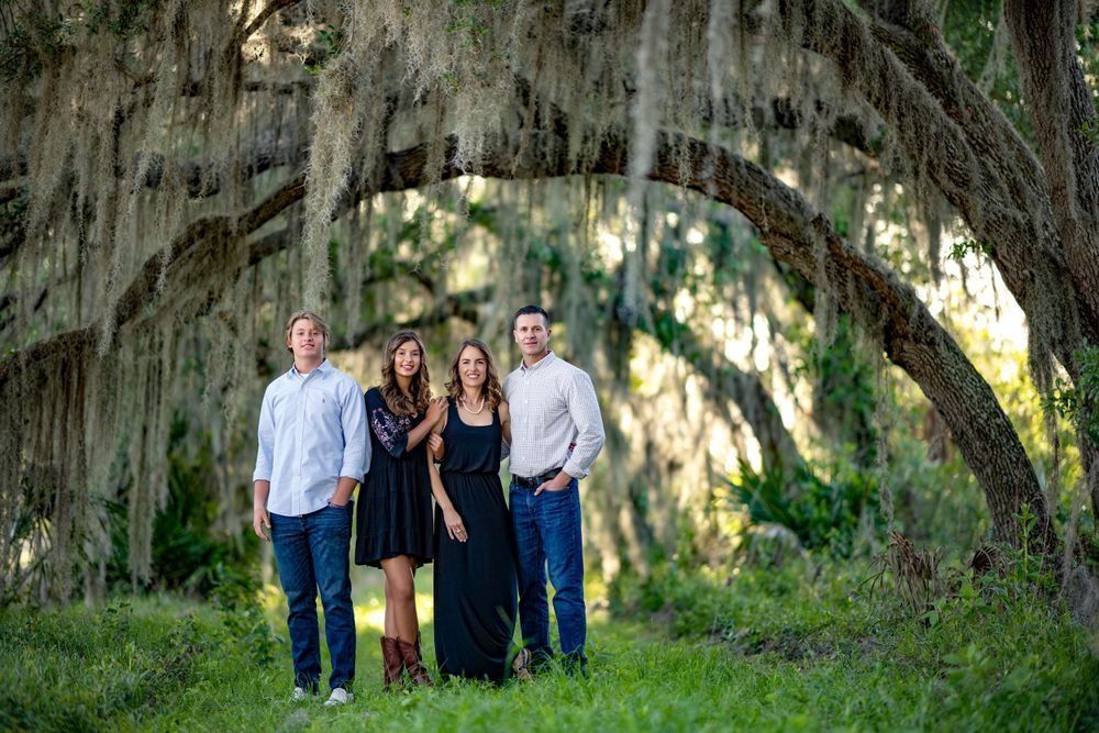 Family posing outdoors under Spanish moss-draped trees. Two women and two men stand in green grass.