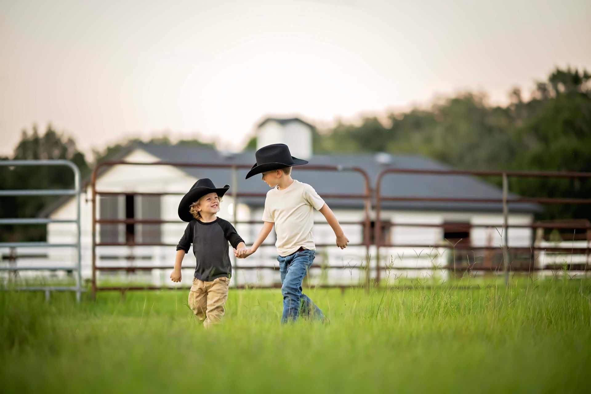 Two young boys in cowboy hats running in a field, holding hands near a fence and house.