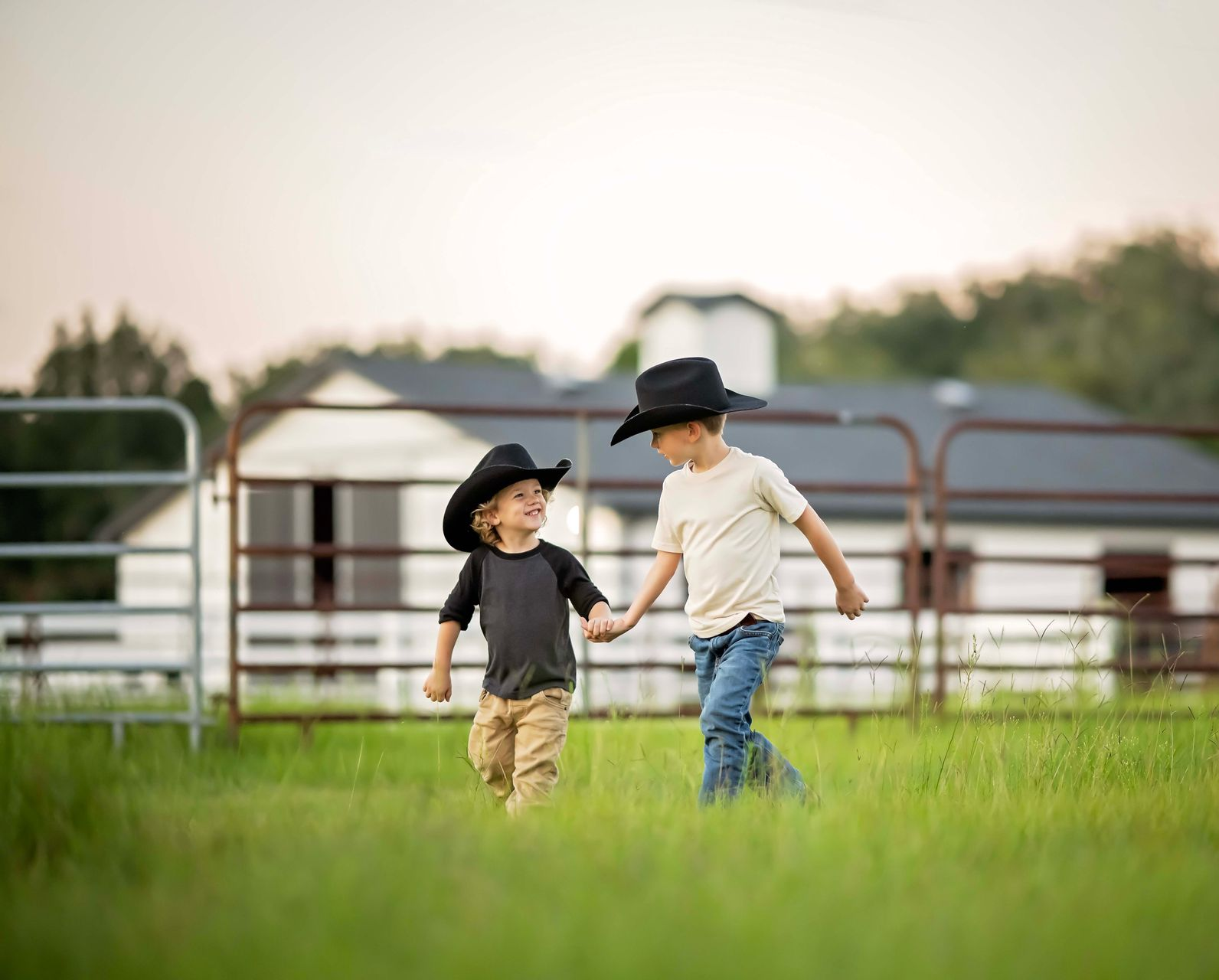 Two young boys in cowboy hats holding hands and walking in a grassy field.