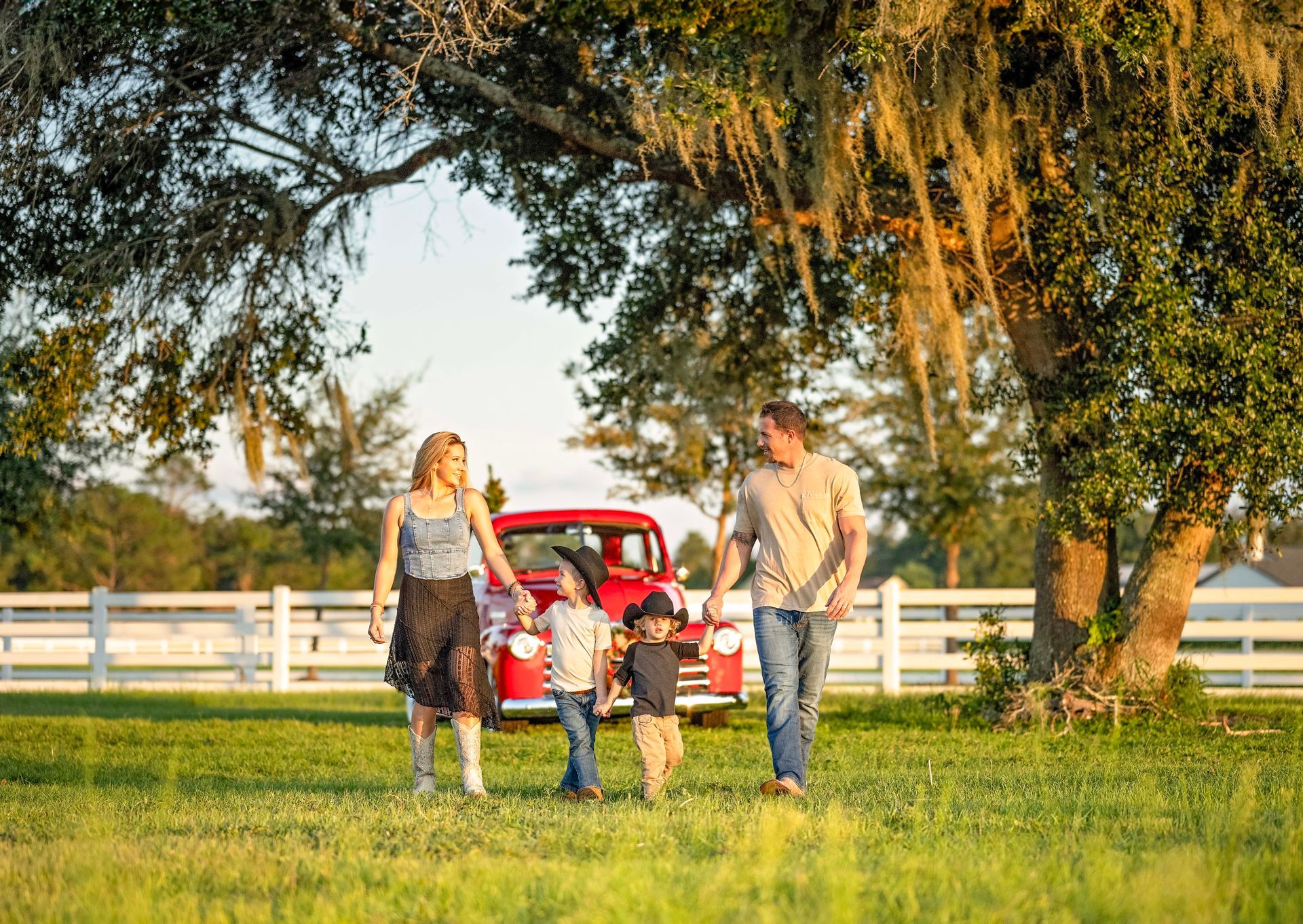 Family walking in a field toward a red vintage truck, under a tree. Sunlight, green grass, and white fence.