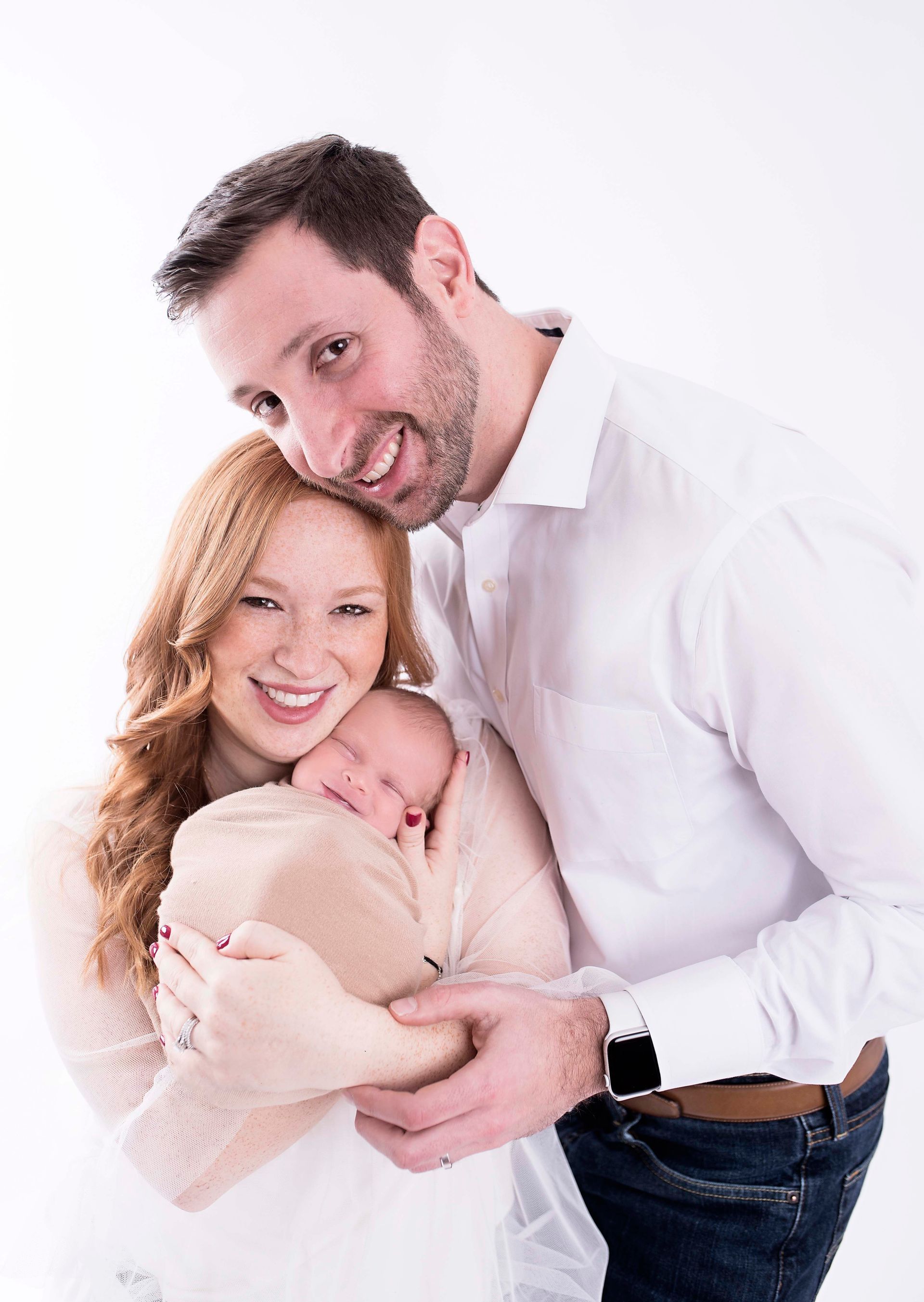 Family of three posing for portrait: mom holding newborn, dad smiling. White backdrop.