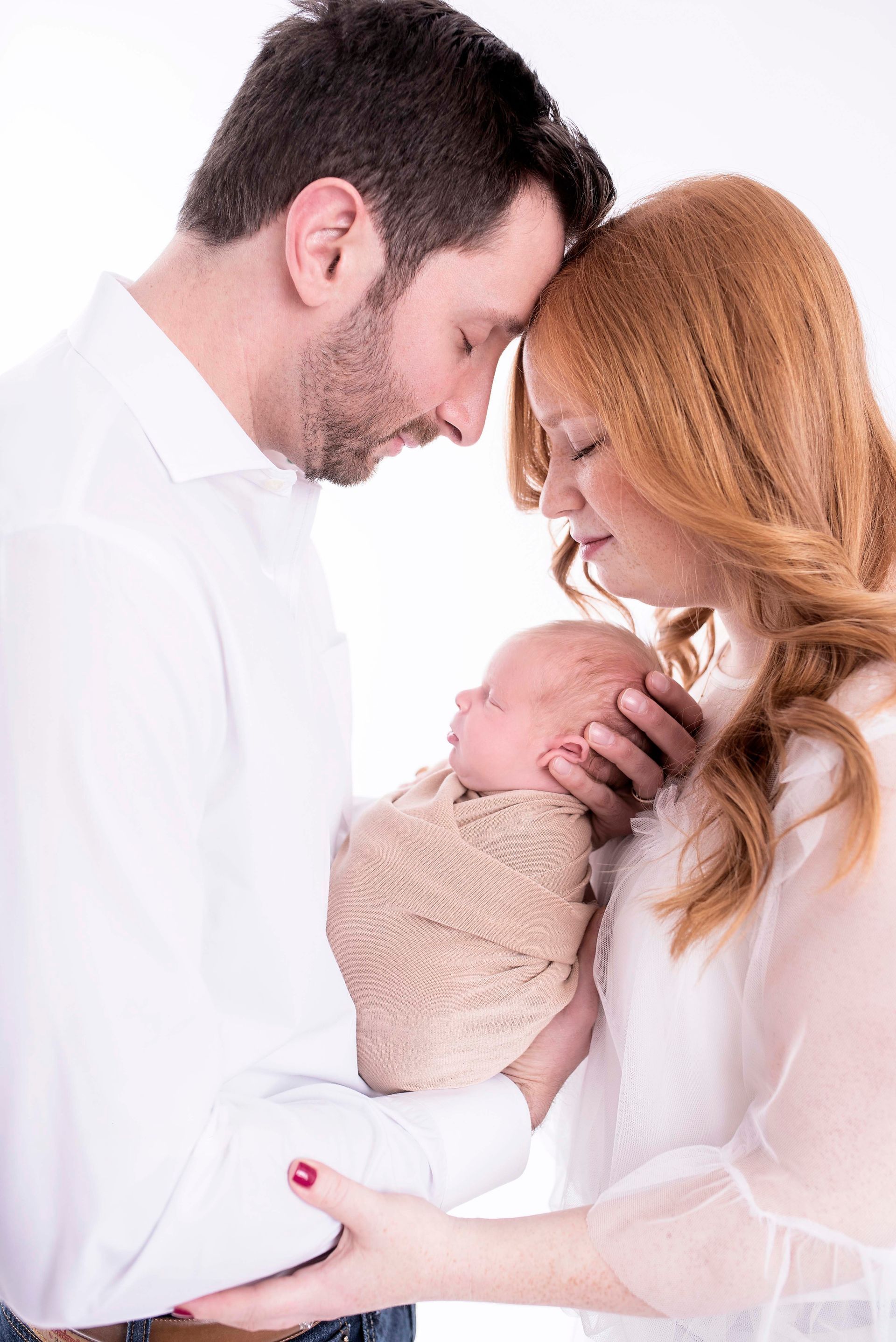 Parents, Caucasian, holding newborn baby wrapped in tan swaddle, heads touching, against white background.