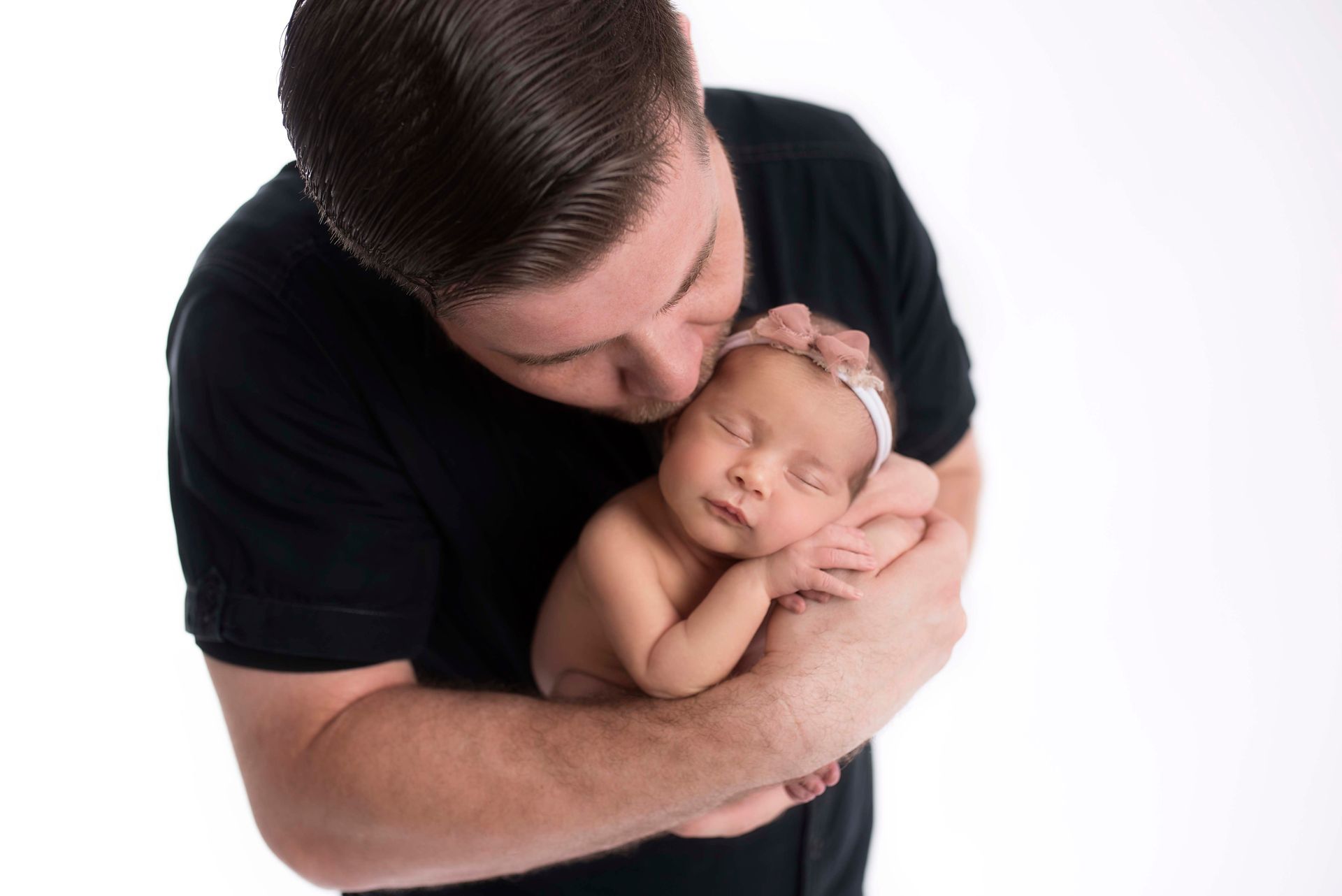 Father kissing a sleeping newborn baby; both are in a bright setting. The man wears black, the baby wears a headband.