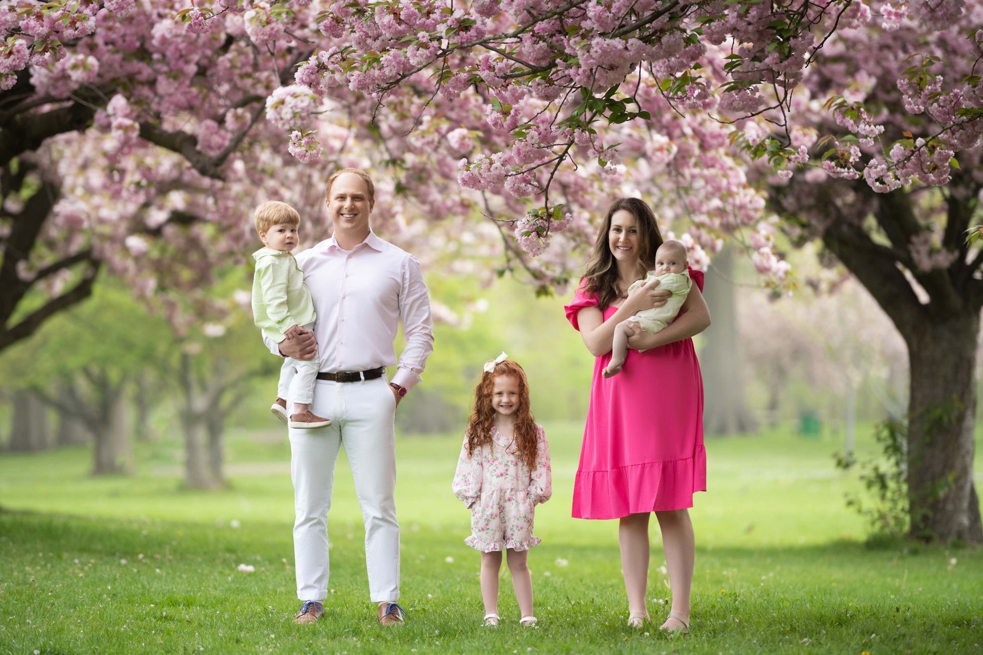 Family of five posing under pink flowering trees in a park; father holding son, mother with baby, daughter standing.