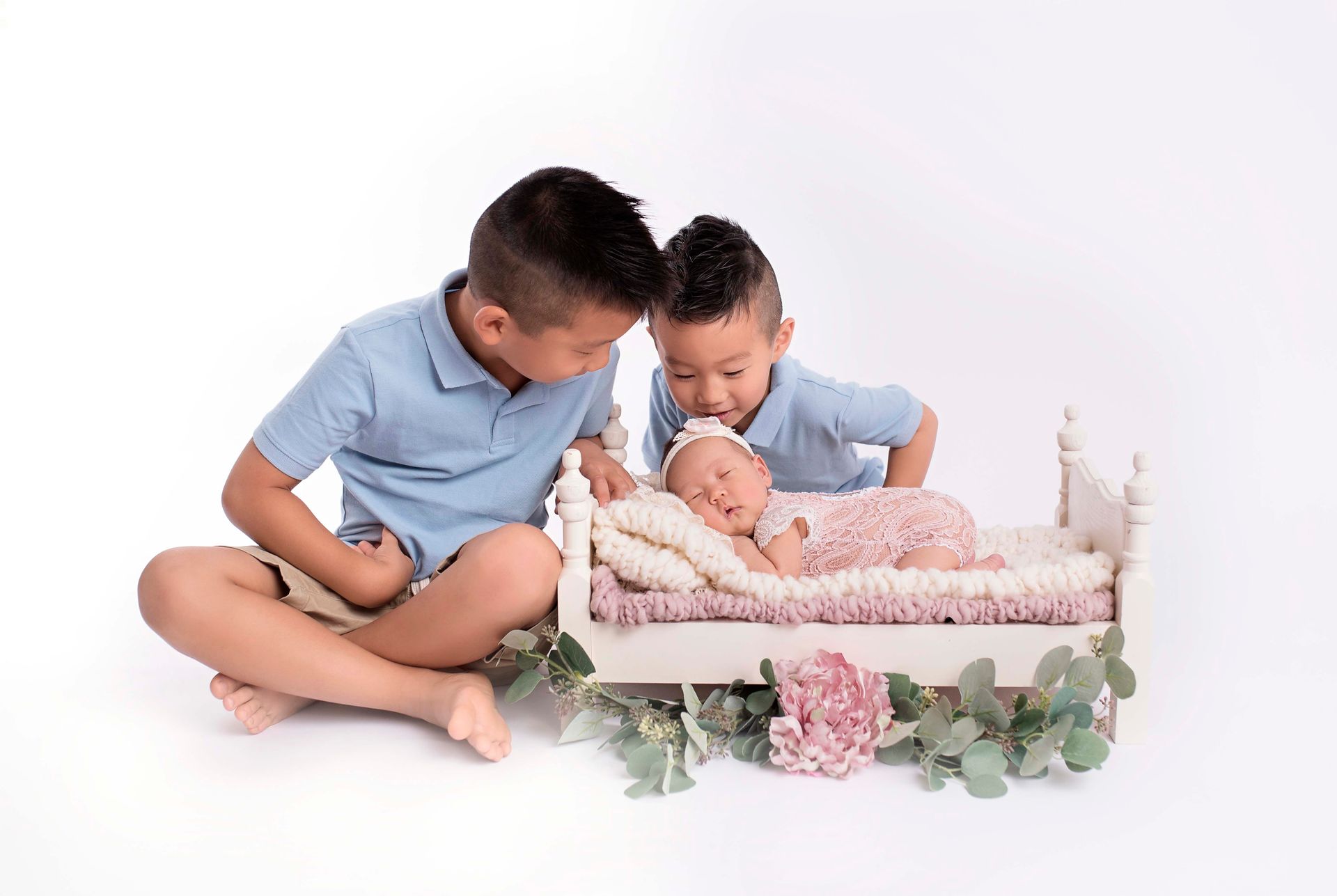 Two boys in blue shirts admire sleeping newborn in small bed with floral accents.