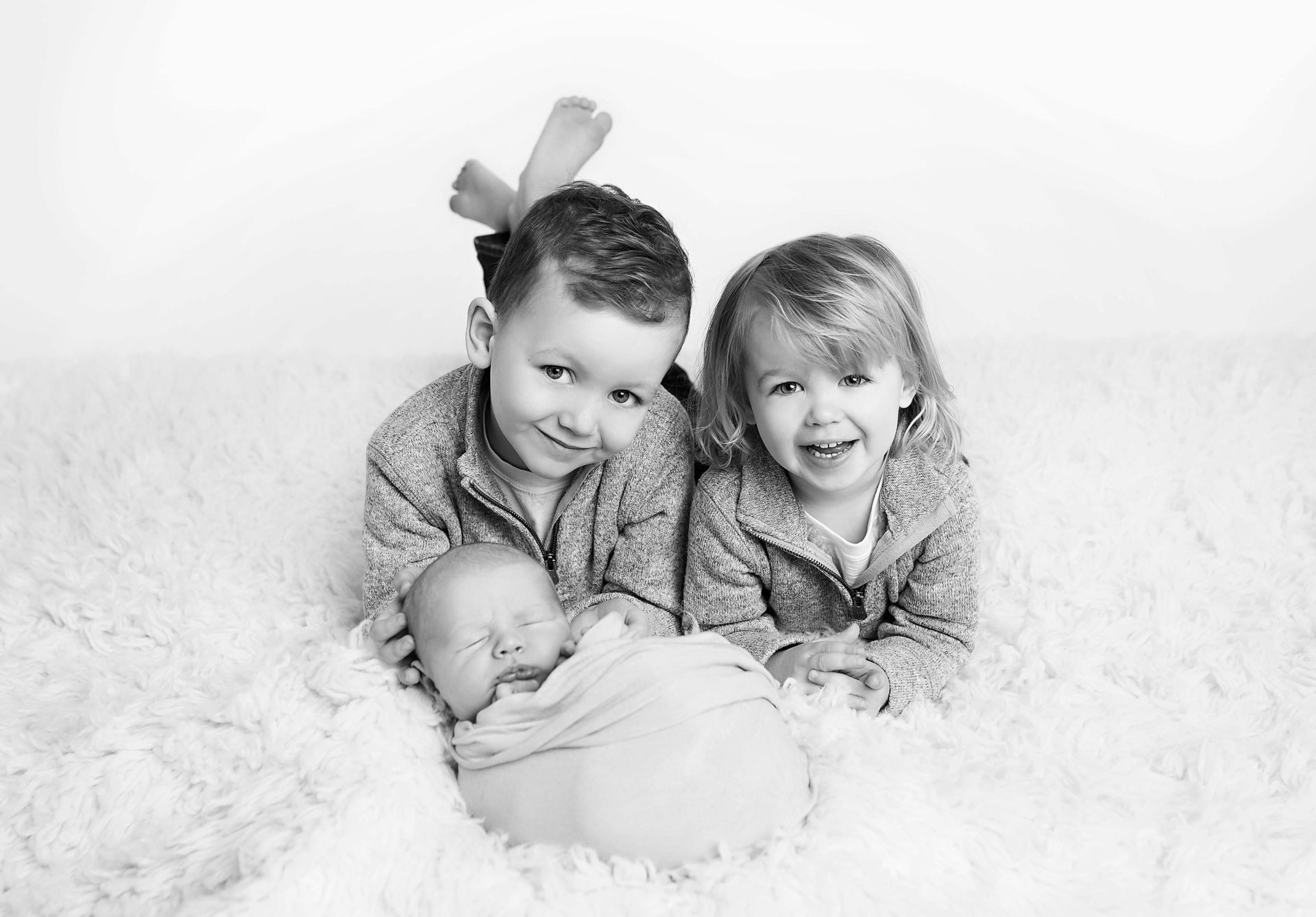 Three siblings, a baby wrapped in a blanket, and two older children smiling, lying on a fluffy white surface.