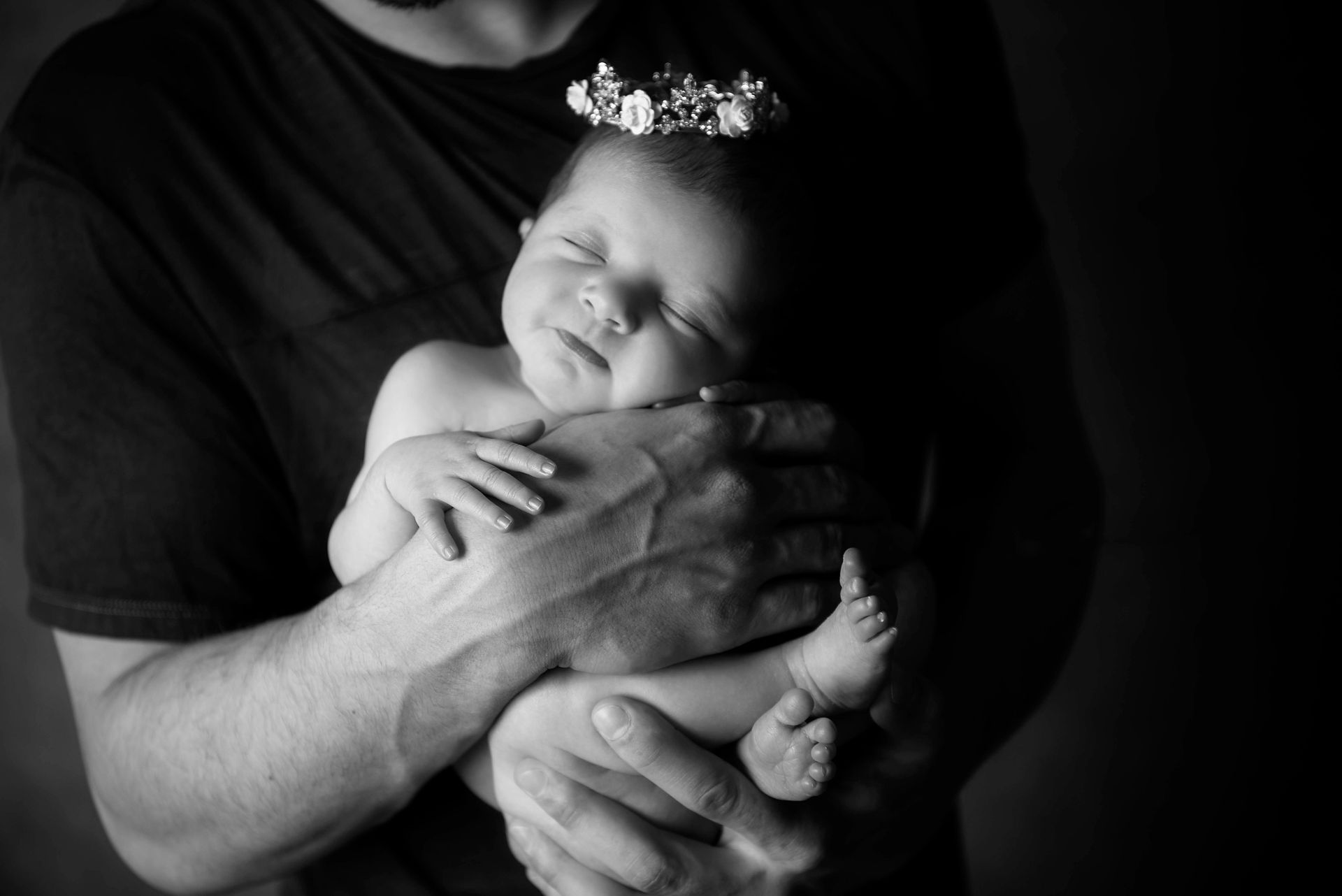 Newborn baby, eyes closed, wearing flower crown, held by person's arms, in black and white.