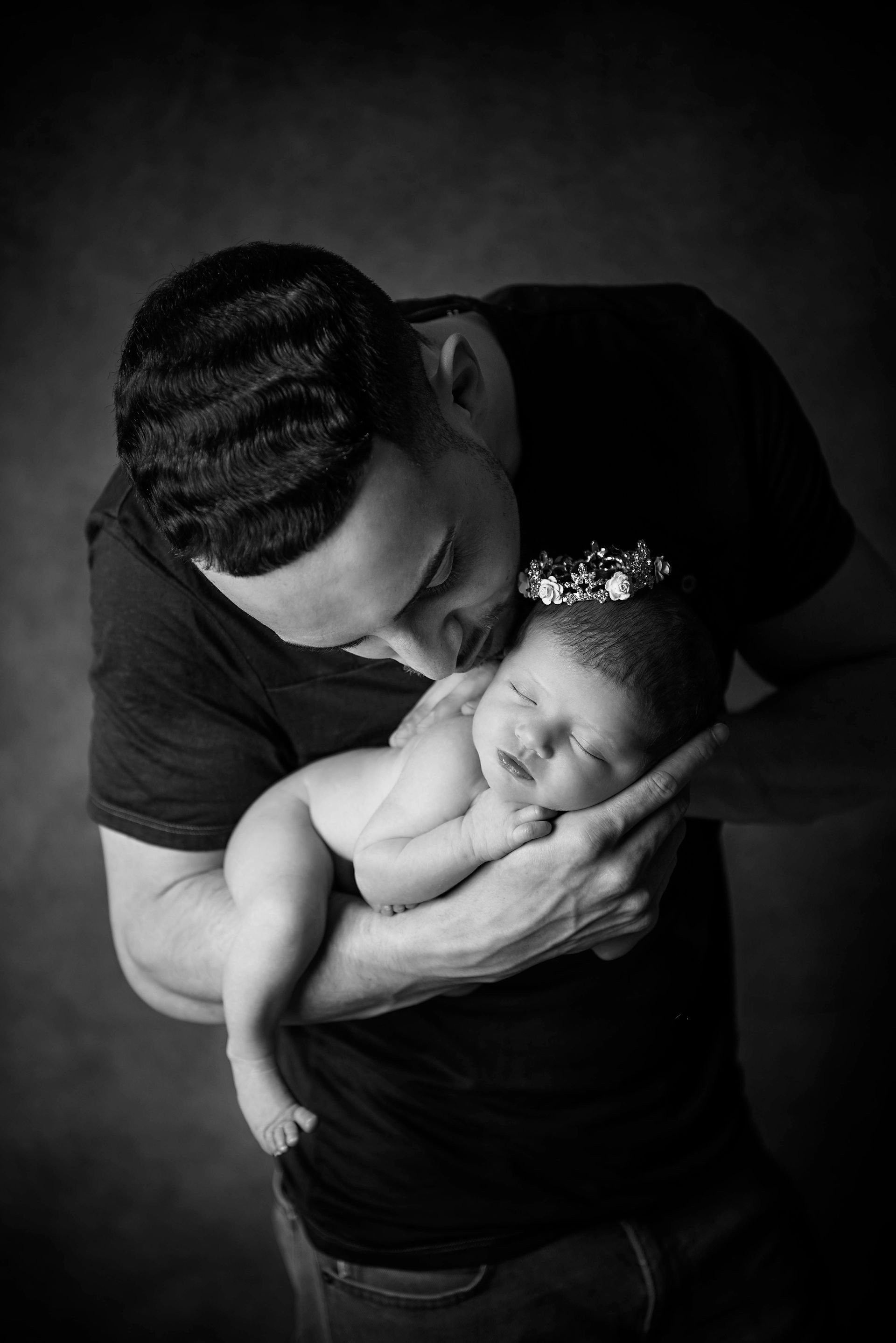 Father holding and kissing sleeping baby, who wears a crown. Black and white.