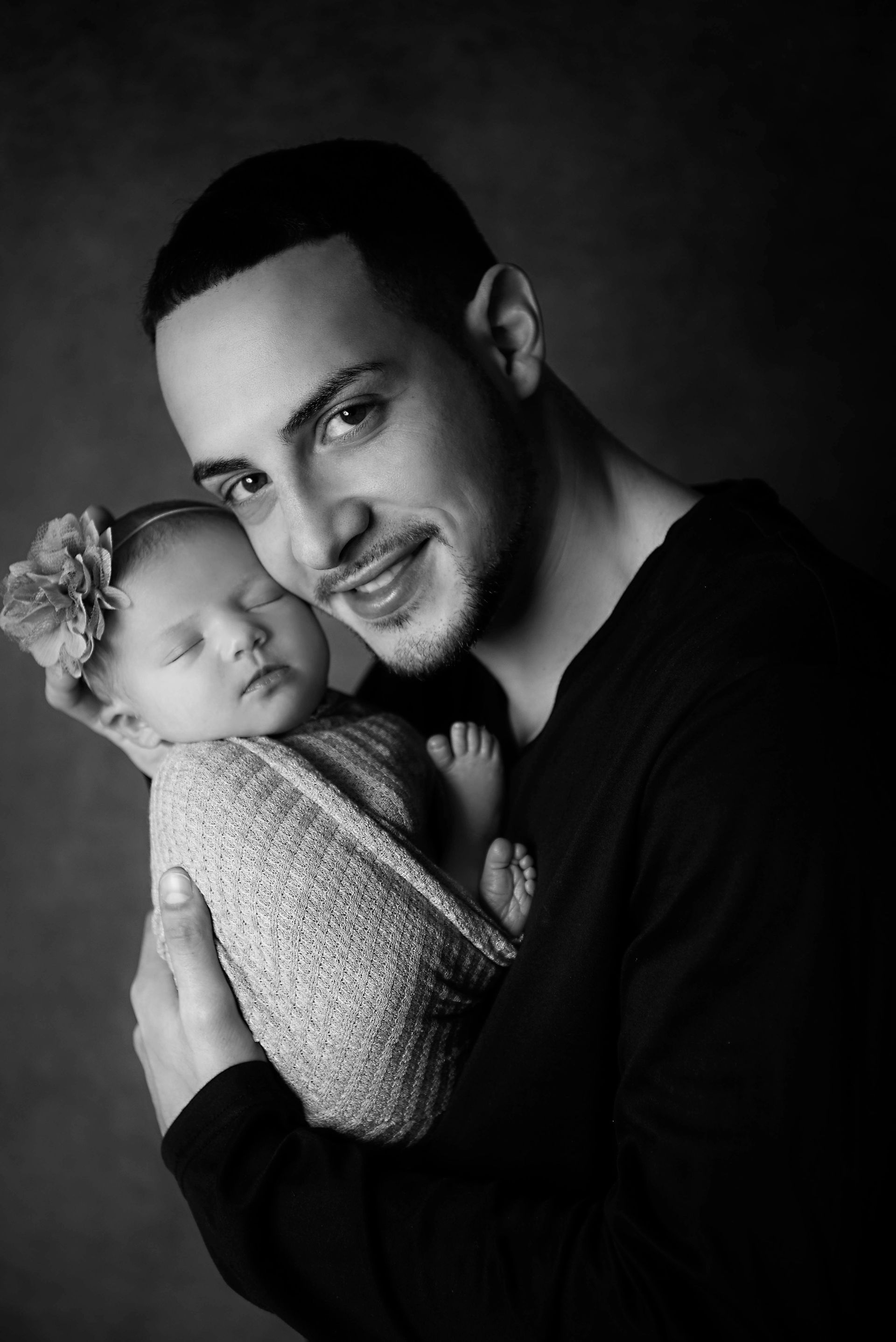 Father holding sleeping newborn, both in black and white. Baby is wrapped in a blanket, father smiles.
