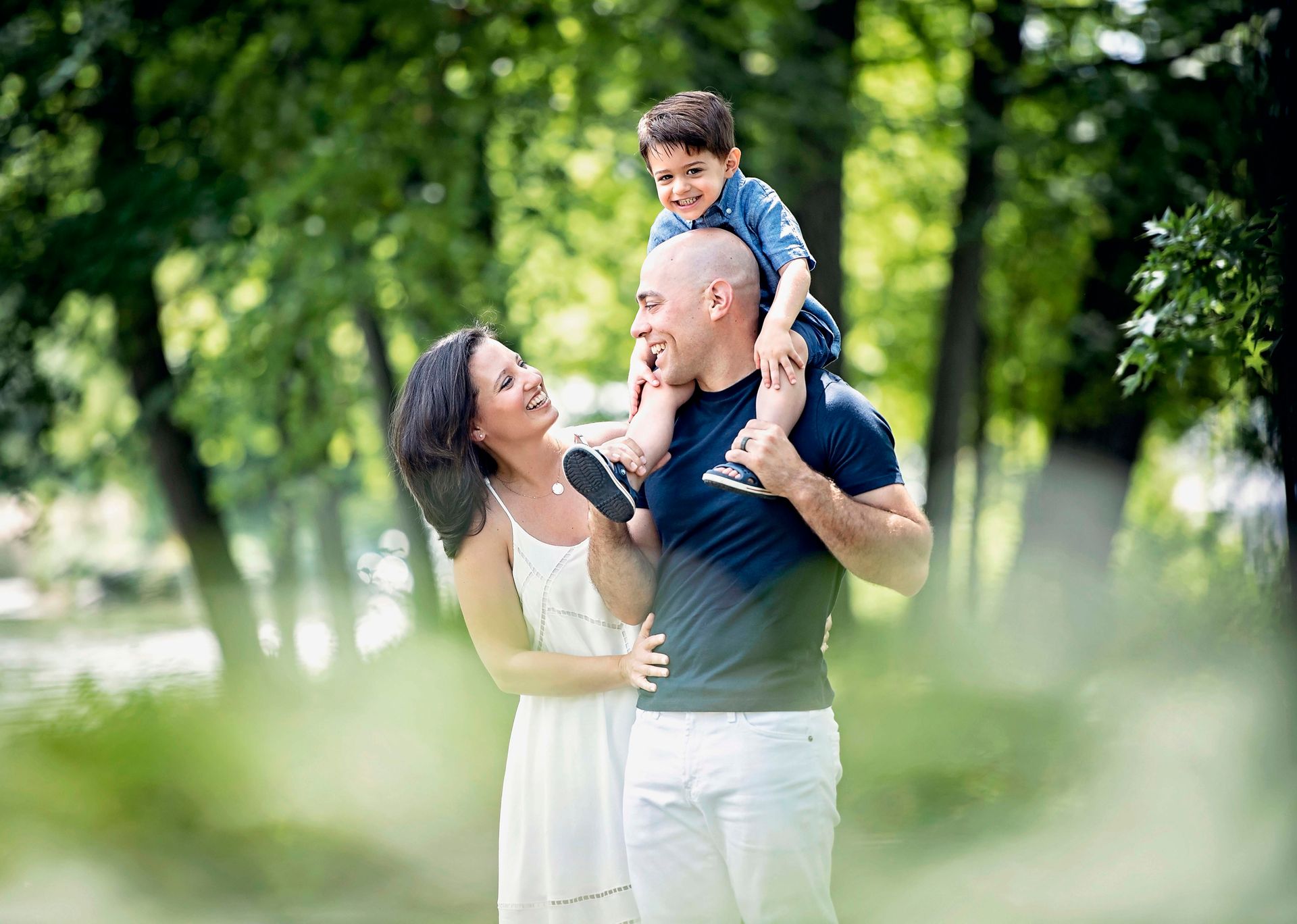 Family of three smiles in a park; child rides on father’s shoulders. Mom wears white, trees in background.
