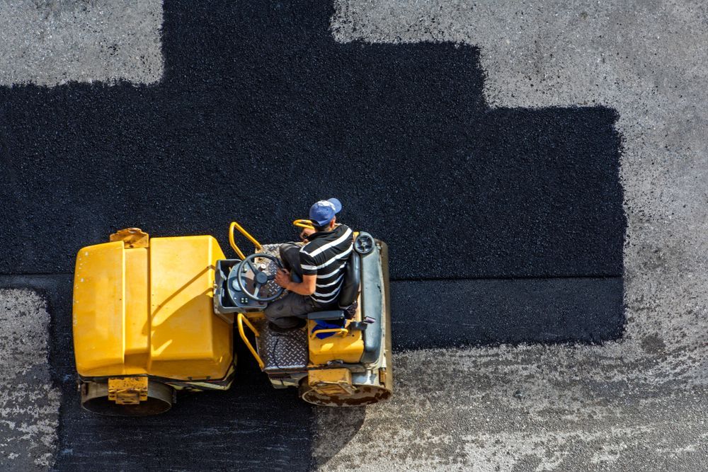 A worker in a yellow road roller paving a dark asphalt surface.