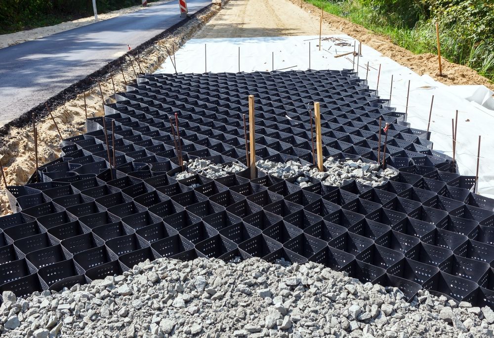 Black plastic ground grid installed over gravel beside a road under construction