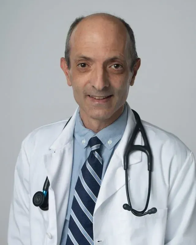 Man wearing a white coat and stethoscope, smiling, looking towards the viewer.