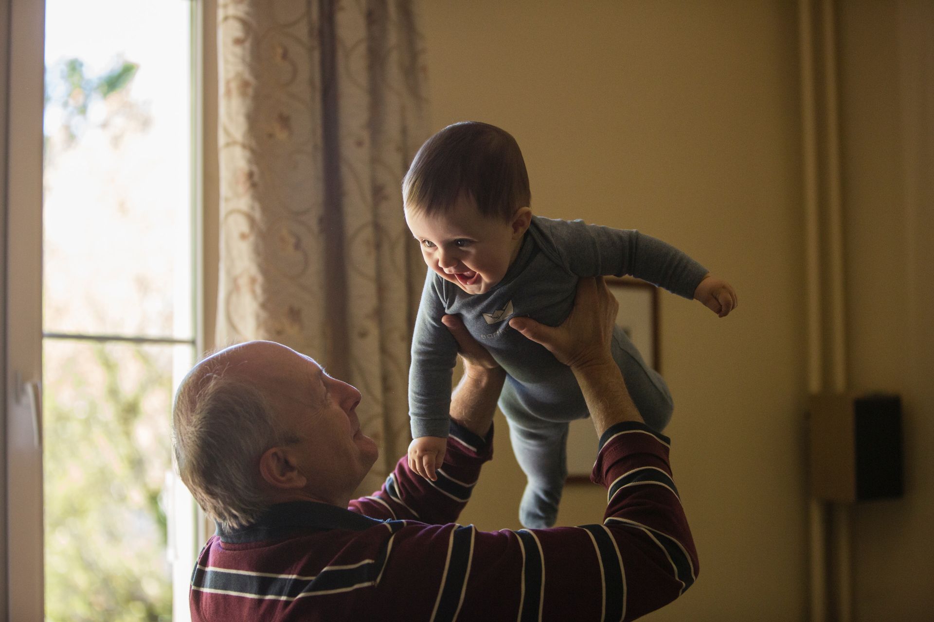 A person holding a baby up in the air indoors near a window, baby smiling.