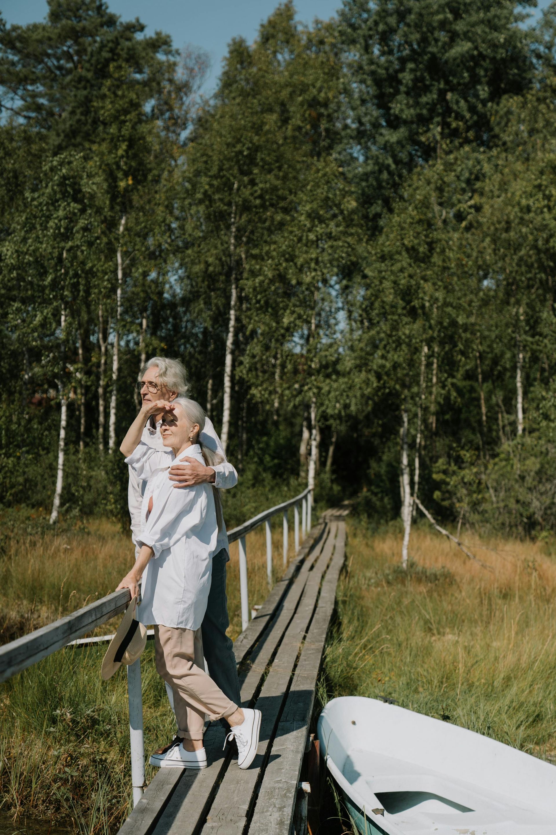 Two people on a wooden pier, looking out. Trees and boat in the background. Sunny day.