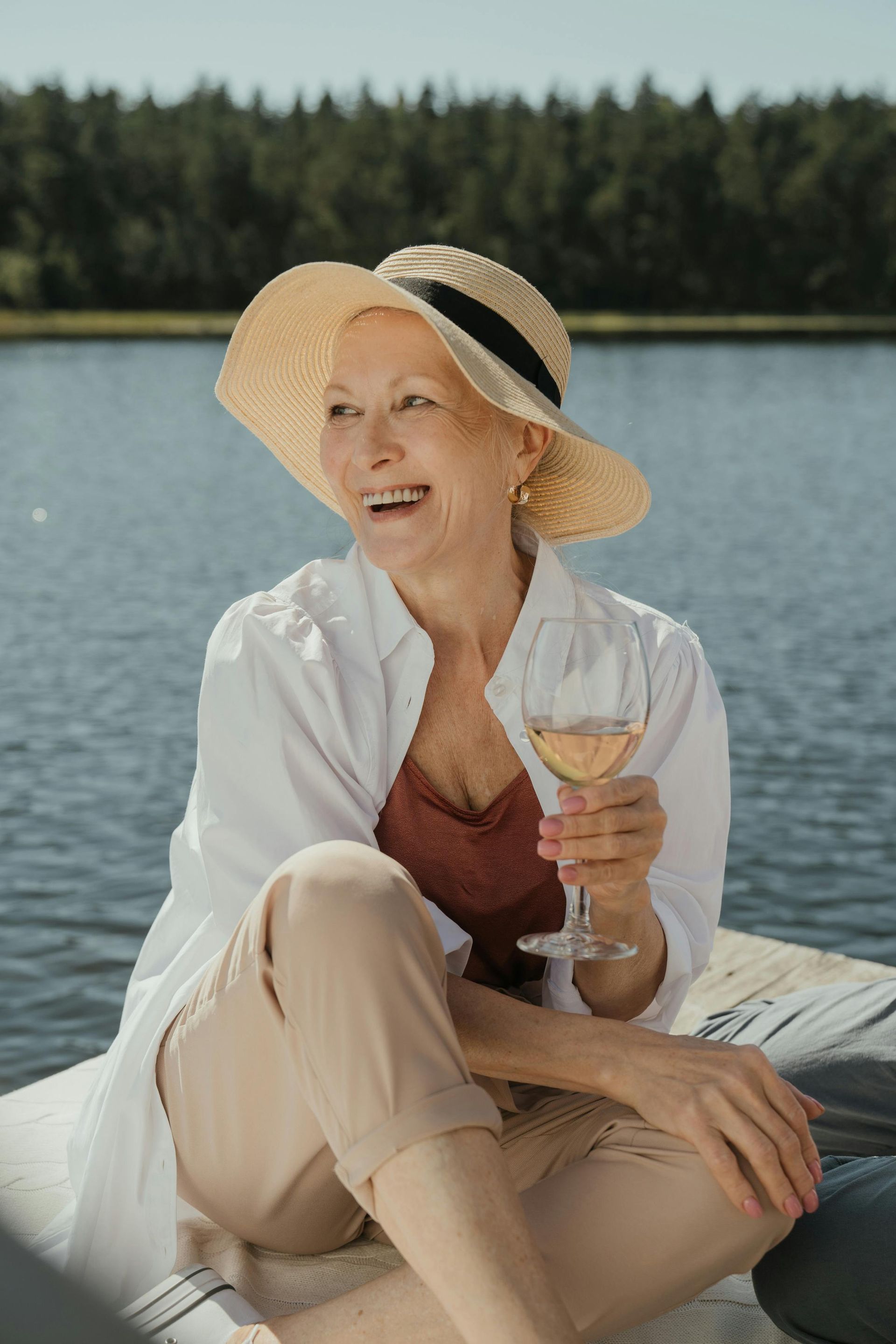 Woman in straw hat smiles, holding wine glass near a lake.