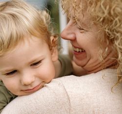 Blonde toddler resting head on grandmother's shoulder, both smiling.
