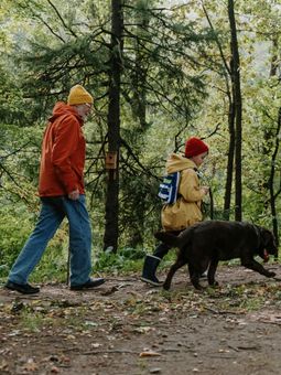 Person in orange jacket and child in yellow raincoat walk in forest with dog.