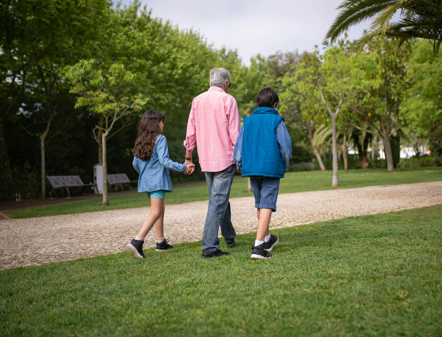 Man walks with two children in a park on a path, holding hands, sunny day.