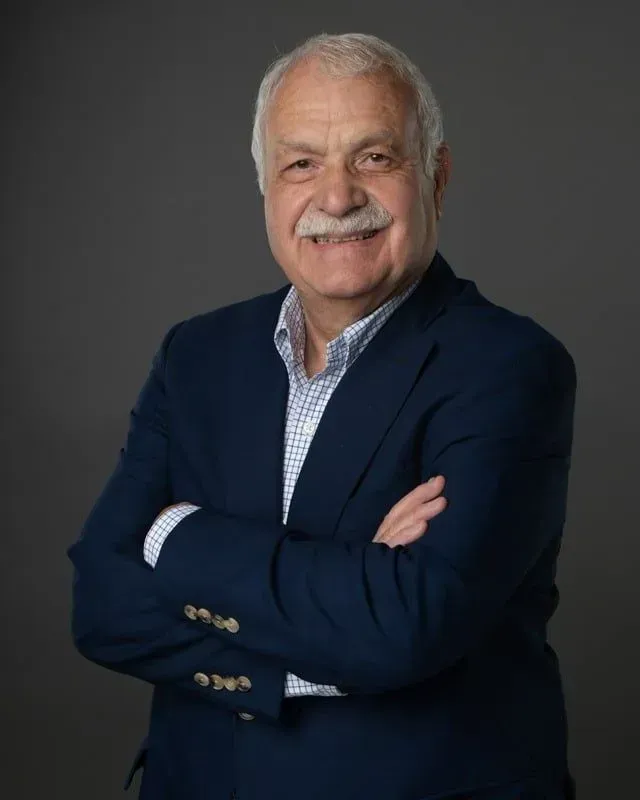 Man with a white mustache, arms crossed, wearing a navy blazer and patterned shirt. Grey background.