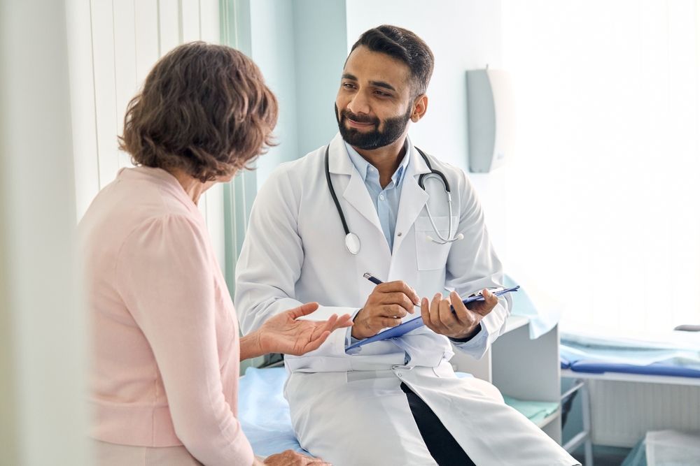 Doctor in white coat speaking with patient, holding a clipboard in a medical room.