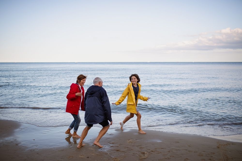 Three people, barefoot, running on a beach, waves in background. One in yellow coat, one in red, one in navy.