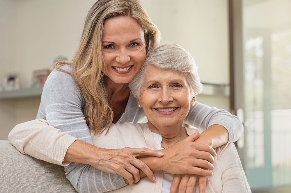 Woman hugs an older woman from behind, both smiling, indoors.