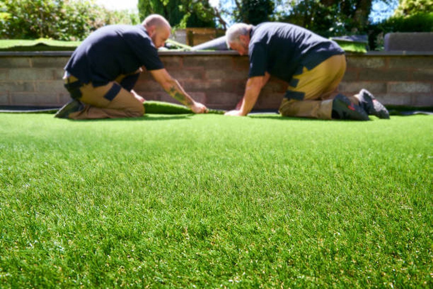 Two men installing artificial turf in a garden, kneeling on the grass.