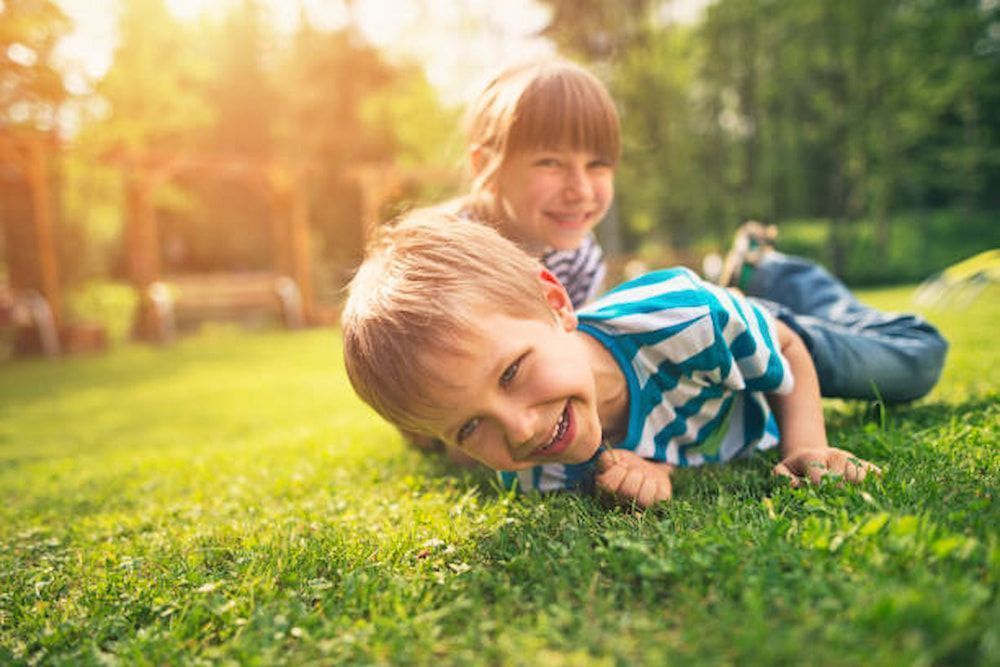 Two children smiling on a grassy lawn with trees in the background, lit by sunlight.