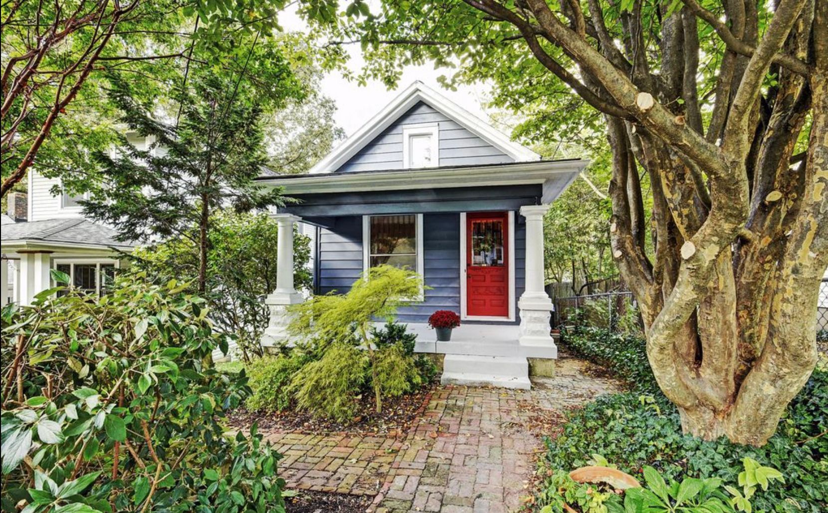 A small blue house with a red door is surrounded by trees and bushes.