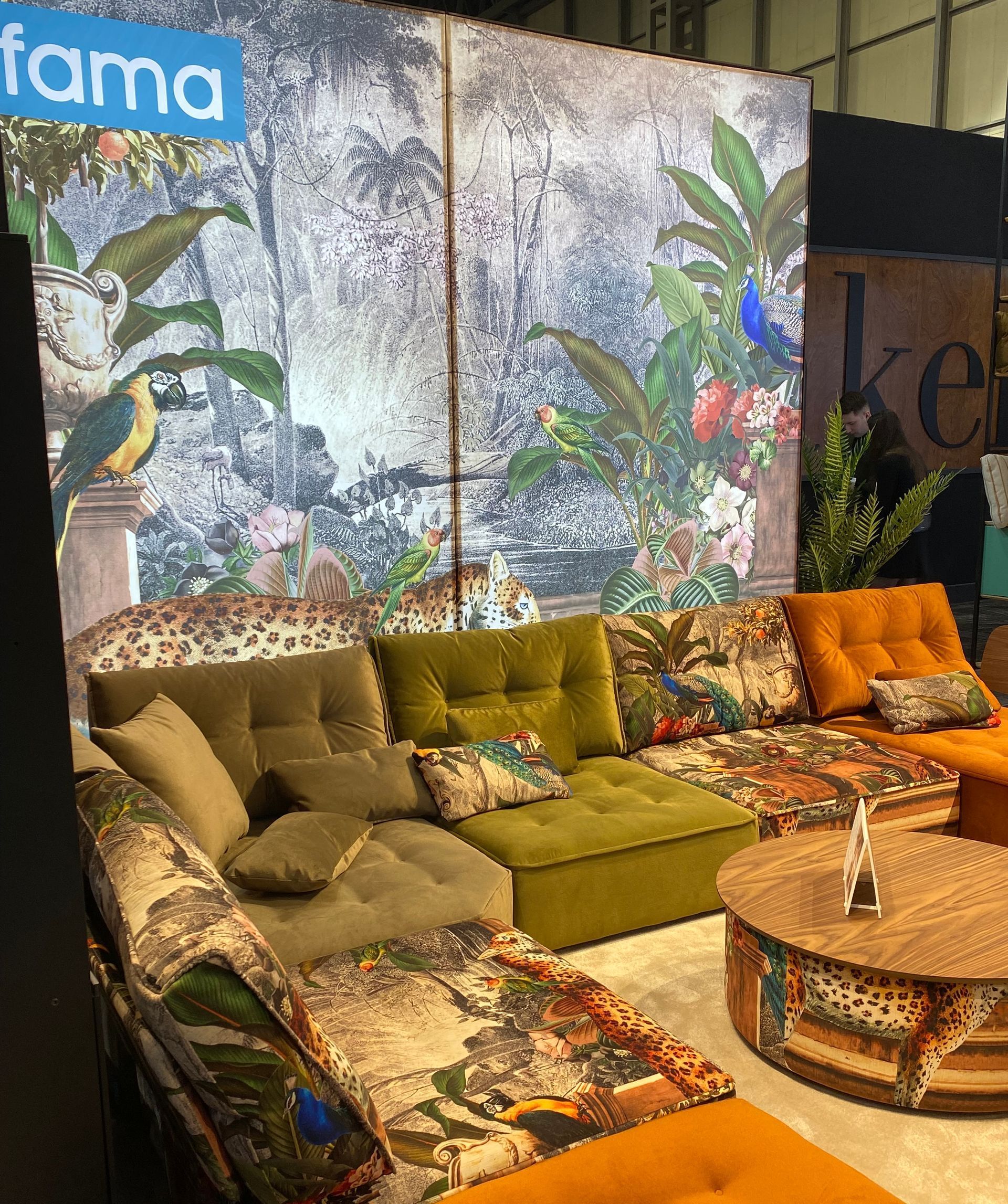 living room display with bespoke green velvet sofa corner system with jungle animal and floral print sections complemented by a wooden coffee table with animal print highlights - all in front of a feature wall with a jungle theme all from 'Fama' at the January Furniture Show 2024, NEC, Birmingham